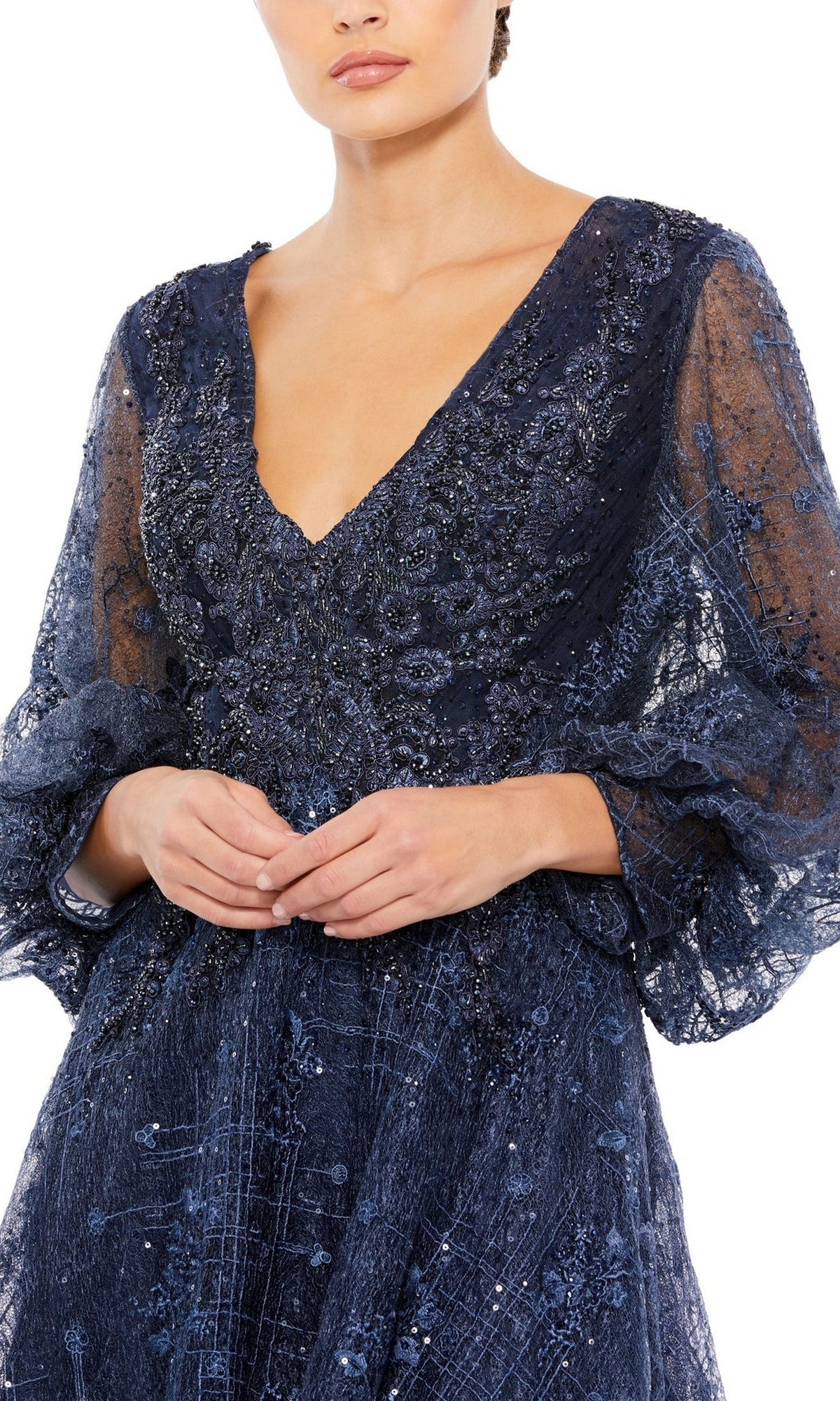 A woman wearing the Mac Duggal Long Formal Dress 20283, a navy blue sheer gown with sequins, embroidery, long sleeves, and a deep V-neckline, stands with her hands clasped in front of her.